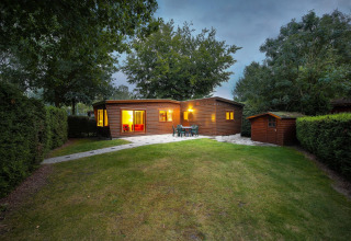 Vista del Cozy Chalet en Bungalowpark Het Verscholen Dorp, Países Bajos, rodeado de césped y árboles al anochecer.