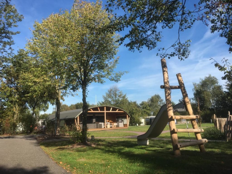 Udendørs lodge og legeplads i ferieparken 't Rheezerwold i Holland med grønne træer og blå himmel.