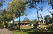 Udendørs lodge og legeplads i ferieparken 't Rheezerwold i Holland med grønne træer og blå himmel.