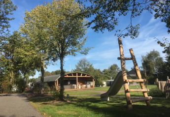 Udendørs lodge og legeplads i ferieparken 't Rheezerwold i Holland med grønne træer og blå himmel.