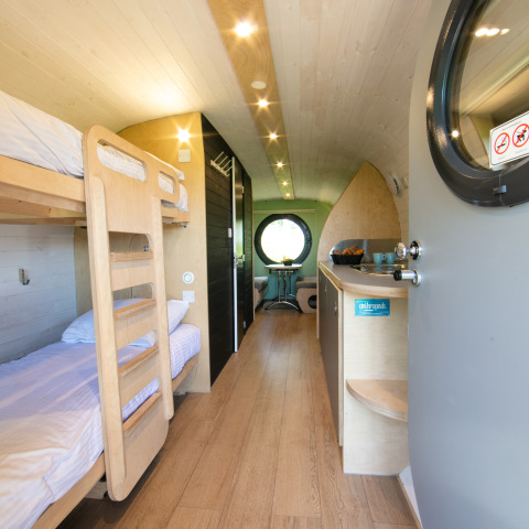 Vista interna di una tiny house con letti a castello, angolo cottura e finestra tonda nei Paesi Bassi.