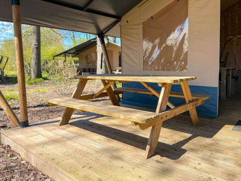 Mesa de picnic de madera sobre una terraza frente a una tienda safari de lujo con sanitario en Camping De Betteld.