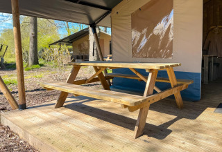 Mesa de picnic de madera sobre una terraza frente a una tienda safari de lujo con sanitario en Camping De Betteld.