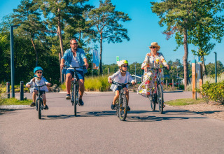 A family enjoys a sunny bike ride together near Kiezel lodge at Zilverstrand in Belgium, surrounded by trees.