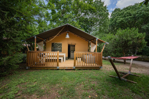 Tienda safari Villatent Nomad con terraza de madera y hamaca en Camping La Clémentine rodeada de árboles.