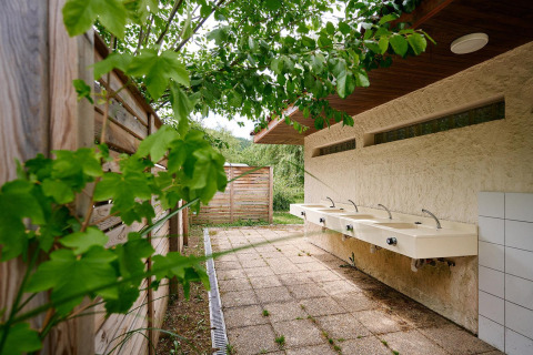 Zona de lavamanos exterior con cuatro pilas bajo techo en Camping de Vaubarlet, rodeada de vegetación.