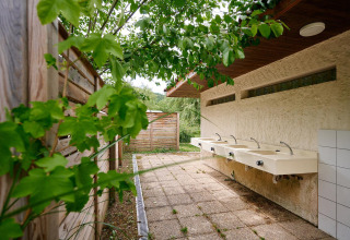 Area lavandini esterna con quattro lavabi sotto una tettoia al Camping de Vaubarlet, circondata da verde.