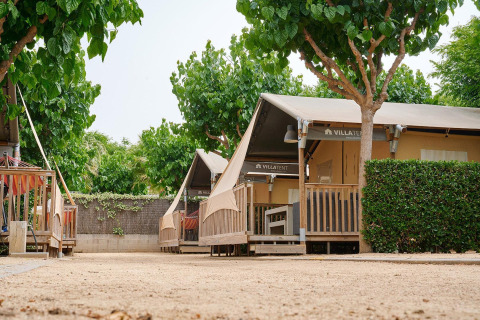 Tienda safari Villatent Outback en Camping La Masia en España, rodeada de árboles y arbustos verdes.