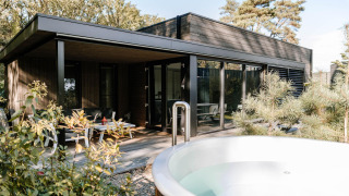 Moderna cabaña de madera con terraza amplia, asientos exteriores y jacuzzi rodeada de árboles verdes.