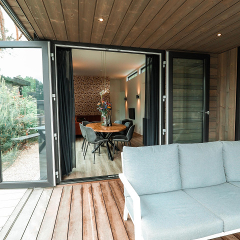 Åben udsigt fra en træterrasse til en moderne lodge med spisebord, stole og stilfuld belysning indendørs.