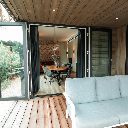 Blick von einer Holzterrasse in eine moderne Lodge mit Essbereich, Sesseln und stilvoller Inneneinrichtung.
