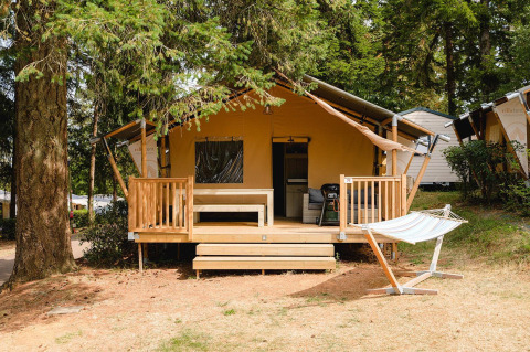 Houten tentlodge met veranda en hangmat bij Villatent Outback op Camping Le Sequoia in Frankrijk, tussen bomen.