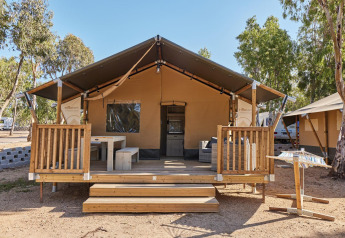 Lodge Villatent Outback con terraza de madera en Camping Village Baia Blu La Tortuga, Italia, rodeado de árboles.
