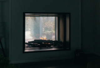 Fireplace inside Veluwe Villa at Holiday park De Boshoek, Netherlands, with garden view through glass.
