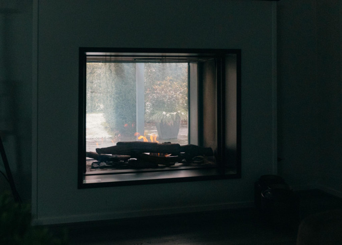 Fireplace inside Veluwe Villa at Holiday park De Boshoek, Netherlands, with garden view through glass.