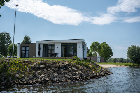 Moderne lodge aan het water genaamd Villa Maritiem, met stenen oever en bomen onder blauwe lucht.