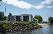 Moderne lodge, Villa Maritiem, gelegen aan het water met rotsen, gras en bomen onder blauwe lucht.
