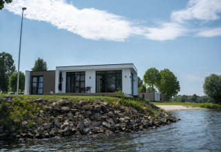 Moderne lodge ved vandkanten med stenstrand og træer i baggrunden under en klar himmel.
