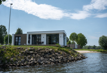 Moderne lodge ved vandkanten med stenstrand og træer i baggrunden under en klar himmel.