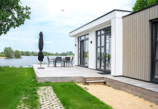 Lodge moderno junto al lago con terraza de madera, mesa de comedor y vistas al agua en Países Bajos.