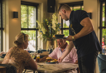 Un camarero sirve comida a dos comensales en una mesa acogedora del restaurante Villa Maritiem.