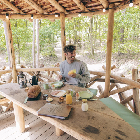 Un hombre desayuna en una mesa rústica de madera en una casa del árbol en Cosy Cabins, bosque de Limburgo, Bélgica.