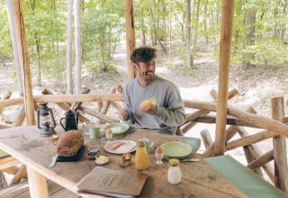 Een man geniet van een ontbijt aan een rustieke houten tafel in een boomhut in Cosy Cabins, Limburgs bos, België.