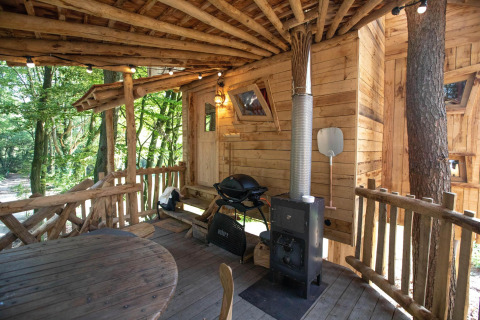 Rustiek houten boomhutterras met een ronde tafel, houtkachel en barbecue, omringd door een weelderig groen bos.