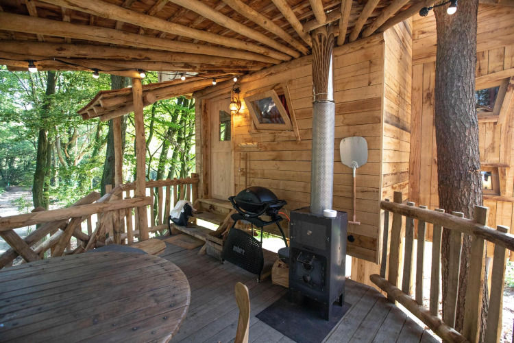 Cubierta rústica de madera para casa de árbol con mesa redonda, estufa de leña y parrilla, rodeada de un frondoso bosque verde.