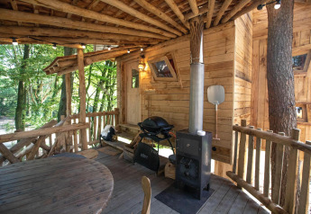 Rustiek houten boomhutdek met ronde tafel, houtkachel en grill, omringd door weelderig groen bos.