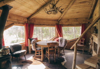 Interior view of a rustic wooden tree house cabin with large windows, dining table, and forest scenery outside.