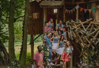 Groep jongvolwassenen in landelijke kleding poseert op de trappen van een versierde boomhut in Limburg, België.