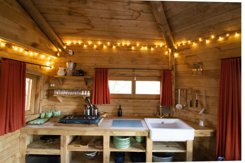 Cuisine rustique en bois dans une cabane dans les arbres avec des lumières de fées, des rideaux rouges et des étagères ouvertes contenant de la vaisselle et des ustensiles.