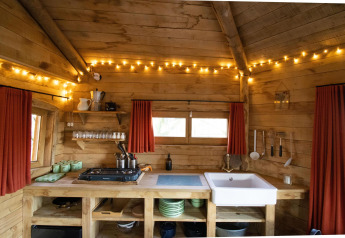 Cucina rustica in legno in una casa sull'albero con luci fiabesche, tende rosse e scaffali aperti con piatti e utensili.