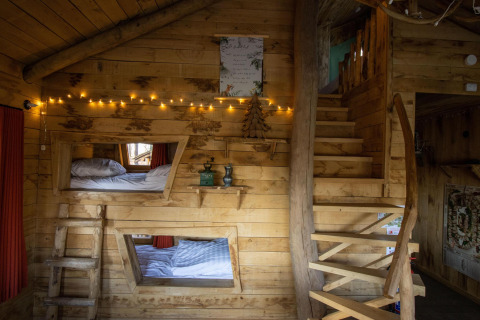 Acogedor interior de casa del árbol de madera con luces de hadas, literas, escaleras rústicas y detalles de madera natural en Bélgica.
