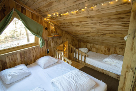 Acogedora habitación de madera en una casa del árbol con luces de cuerda, dos camas y una ventana con cortinas verdes en Bélgica.