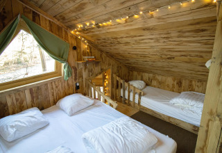 Gezellige houten boomhut slaapkamer met koordverlichting, twee bedden en een raam met groene gordijnen in België.