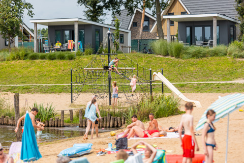 Parque vacacional en la playa con niños jugando en la estructura y gente relajándose frente a cabañas.