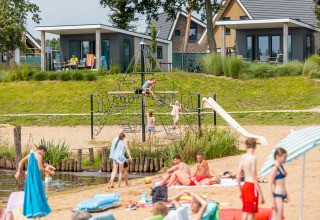 Parque vacacional en la playa con niños jugando en la estructura y gente relajándose frente a cabañas.