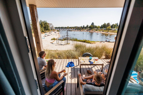 Zicht vanuit een Beach lodge in Vakantiepark Leukermeer, Nederland, met mensen op het terras bij het meer.