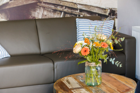 Sala con sofá gris, cojines a rayas y ramo de flores en jarrón de vidrio en Beach lodge en los Países Bajos.