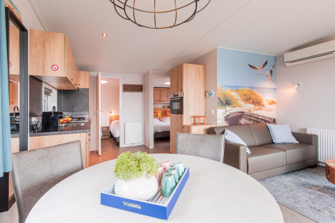 Lyst og moderne strandhytteinteriør med opholdsstue, køkken og strandtema-vægmaleri i Vakantiepark Leukermeer.