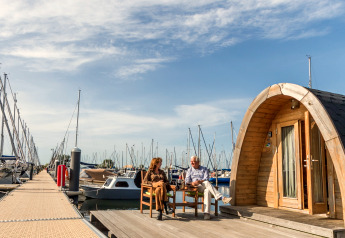Two people sit outside a tiny house named Water bubbles at Marina Parcs Almere, surrounded by boats.