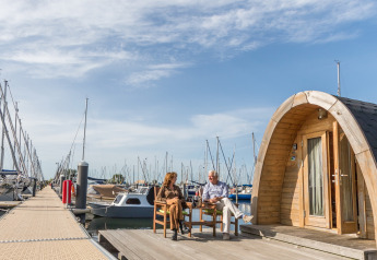 Two people sit outside a tiny house named Water bubbles at Marina Parcs Almere, surrounded by boats.