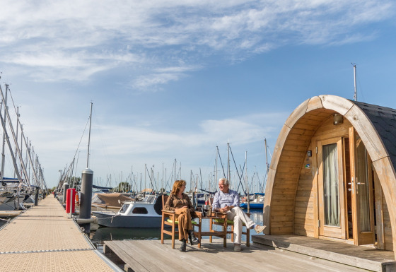 Deux personnes assises devant une tiny house Water bubbles à Marina Parcs Almere, entourées de bateaux.