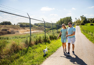 Couple promenant leur chien sous un soleil éclatant près d’une clôture à Biebosch - Sunshower, Resort Mooi Bemelen, Pays-Bas.