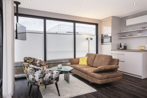 Moderne woonkamer op een woonboot in Offingawier met grote ramen, bruine zetel en bloemige stoelen.