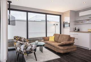 Moderne woonkamer op een woonboot in Offingawier met grote ramen, bruine zetel en bloemige stoelen.