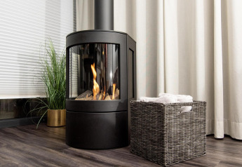 Chimenea moderna junto a una cesta de mimbre y planta verde en casa flotante con cortinas y suelo de madera.