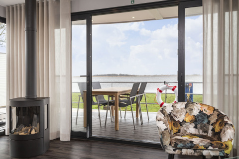 Salón de casa flotante con chimenea, silla floral y terraza con vistas al agua en Friesland, Países Bajos.